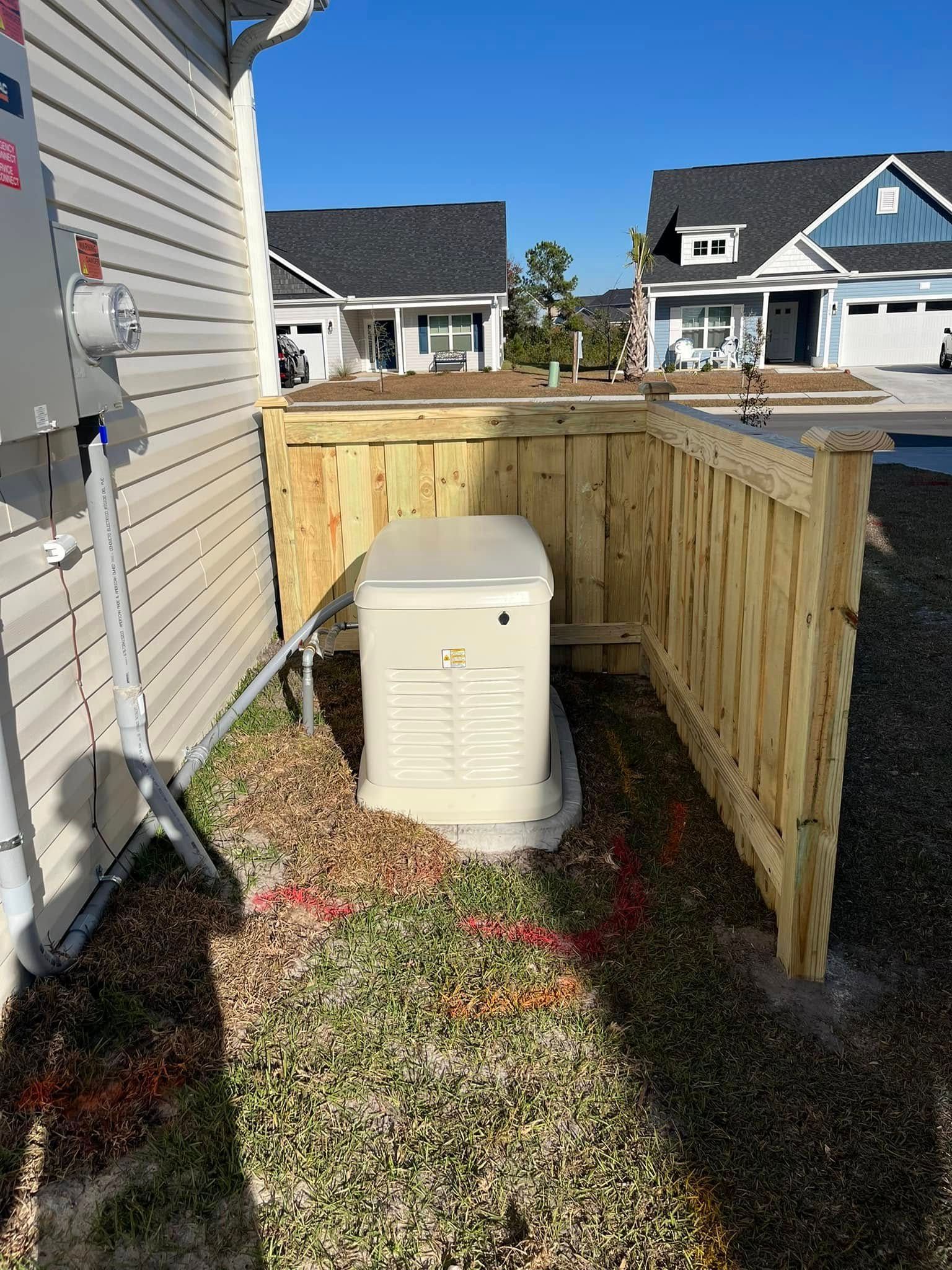 A generator is sitting under a wooden fence next to a house.