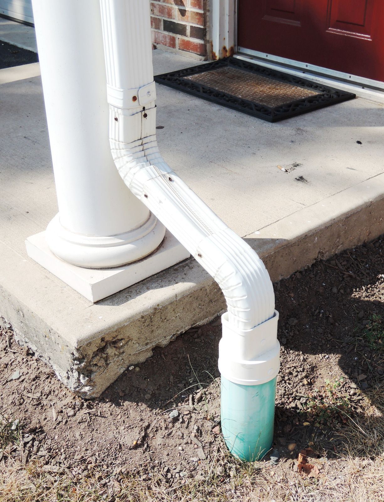 A white drainpipe is connected to a green pipe on the side of a porch.
