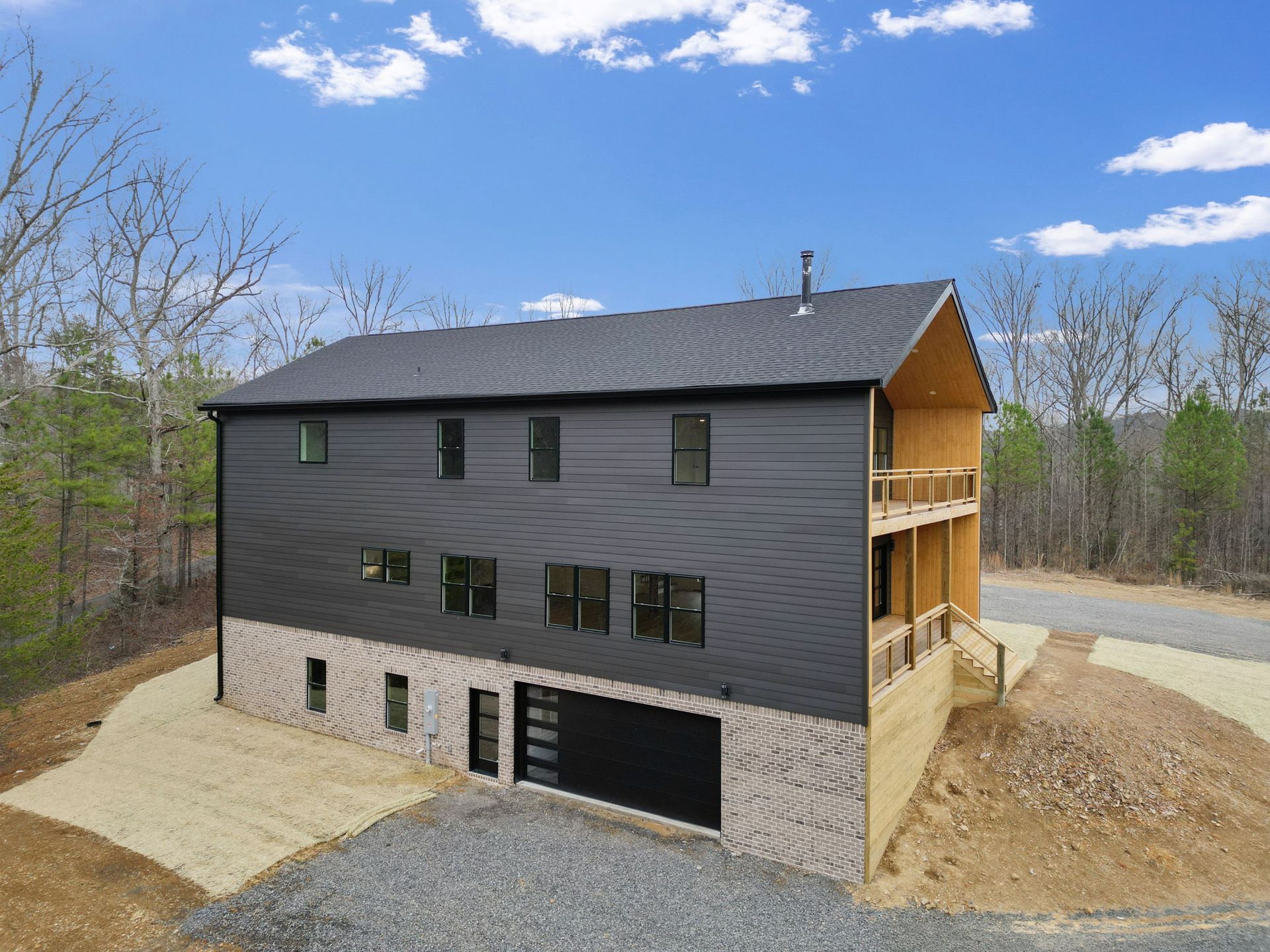 Modern black siding home with brick foundation, covered wood porch, and three-story layout in wooded rural setting.