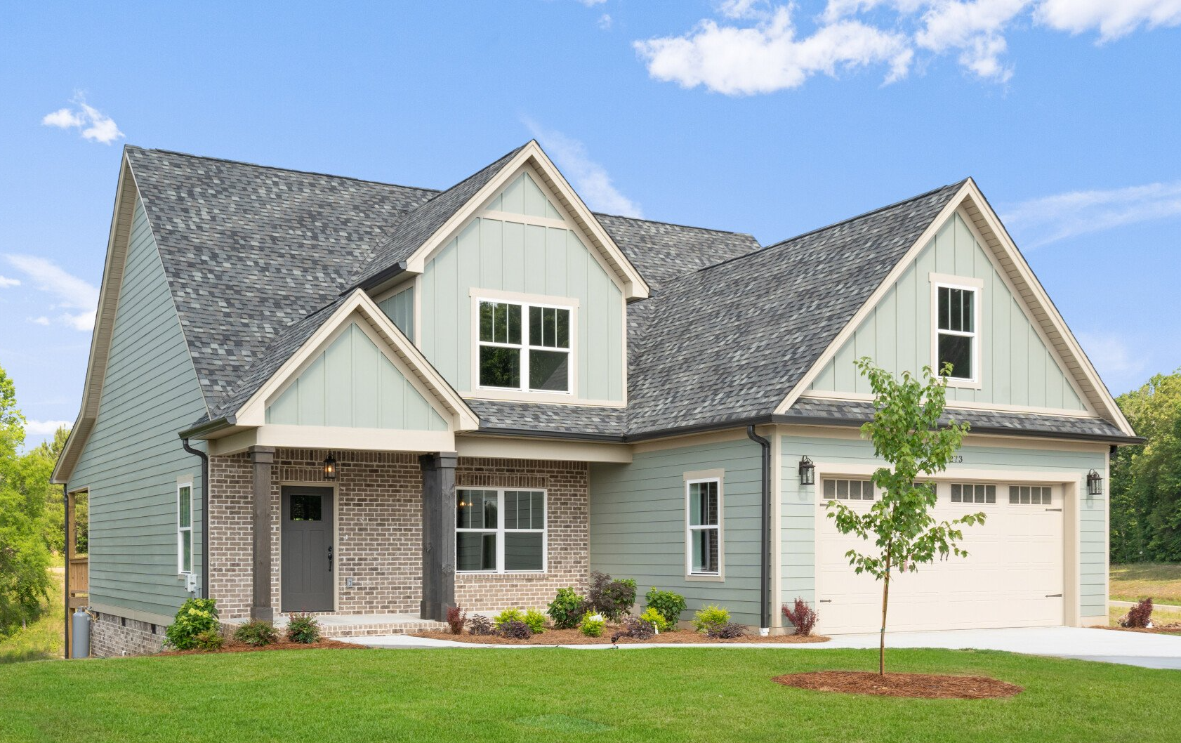 Modern craftsman home with green siding, brick accents, gable roof, and two-car garage in a landscaped suburban setting.