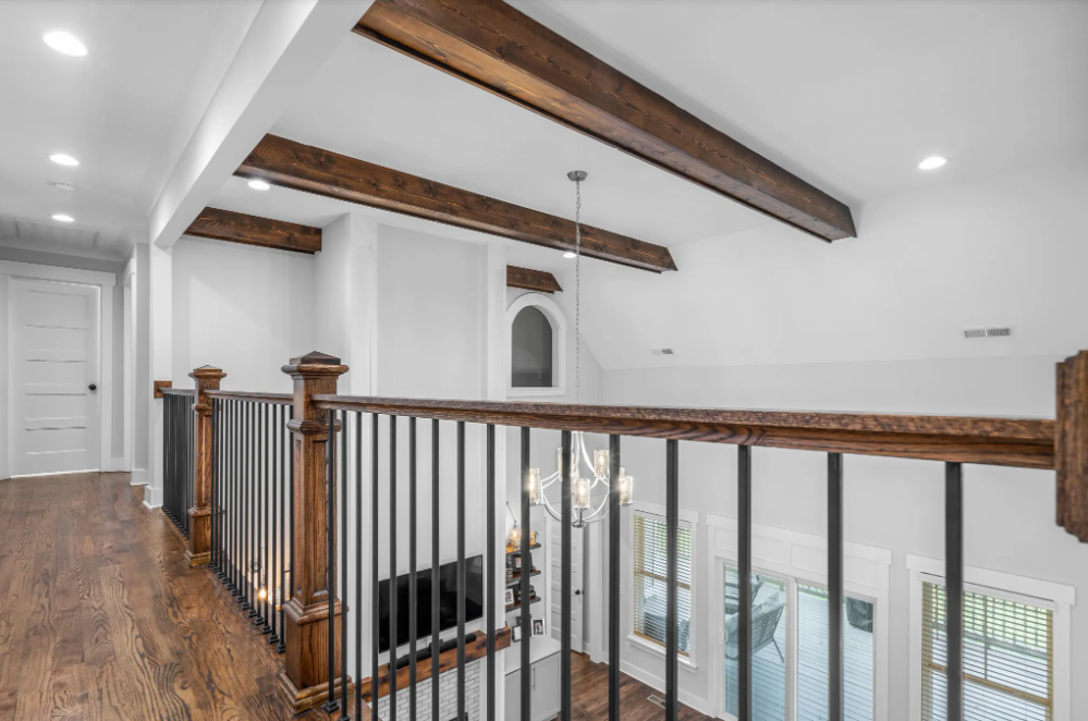 Second-floor hallway overlooking a grand living room with exposed wood ceiling beams, iron and wood railing, hardwood floors, and chandelier lighting.