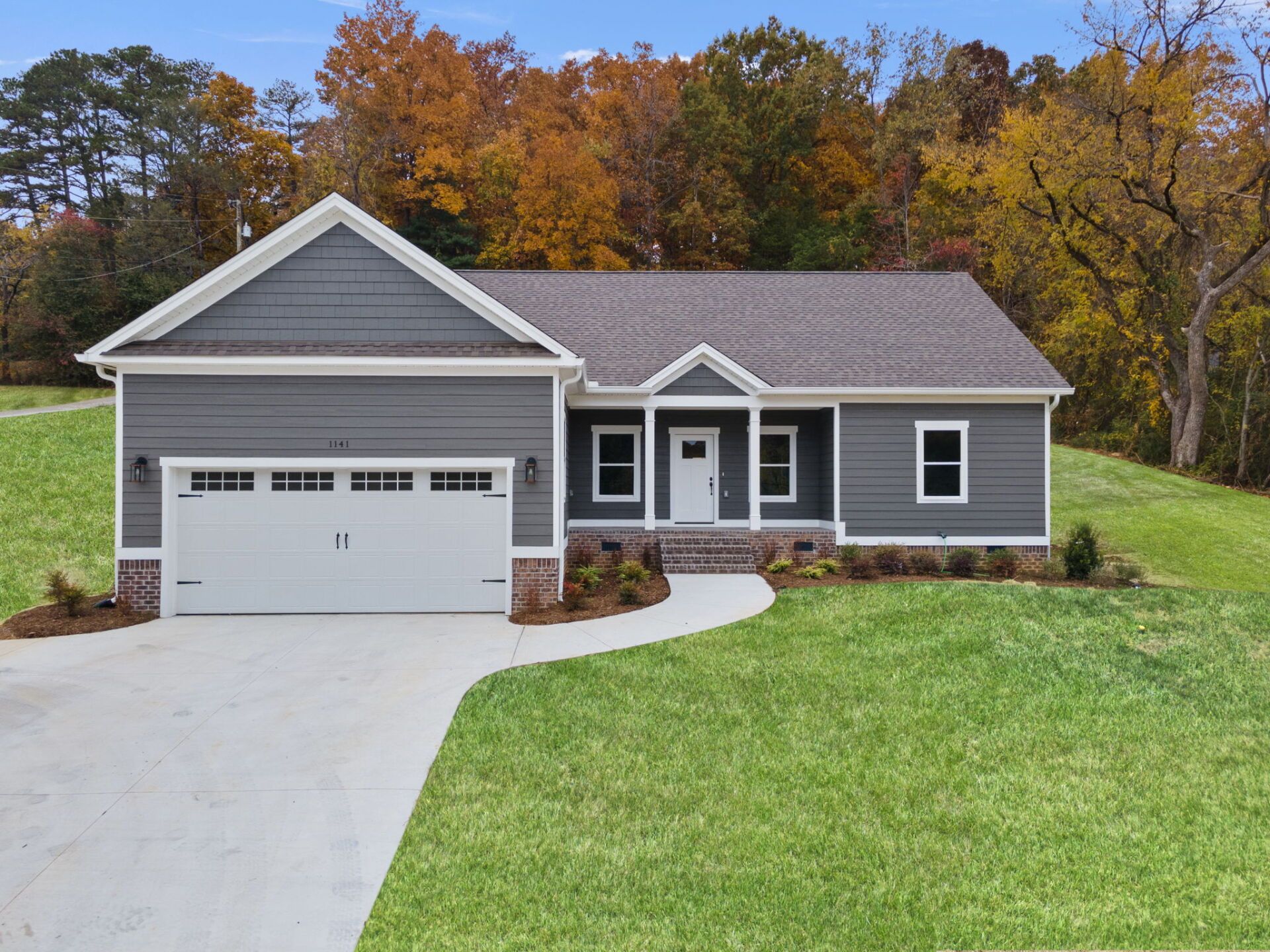 Gray ranch-style home with white trim, two-car garage, and covered front porch on a spacious green lawn with fall trees in the background.