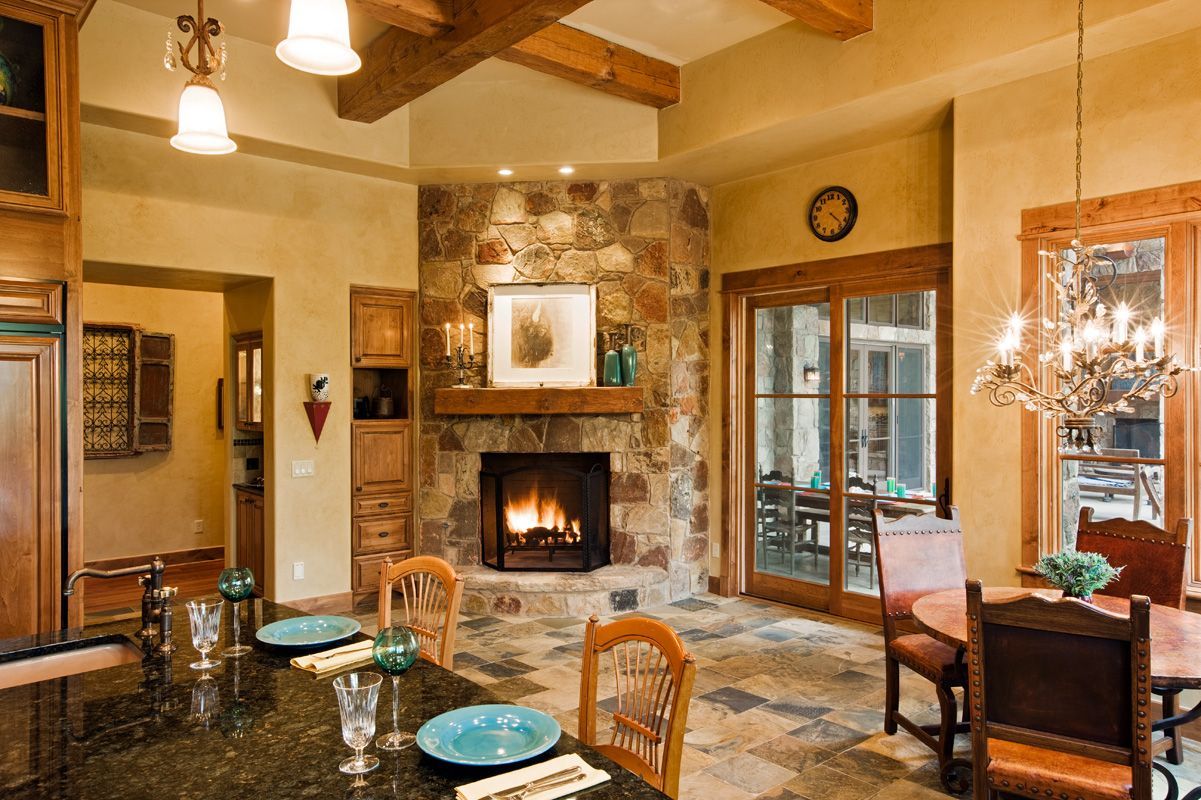 Cozy kitchen with stone fireplace, wooden beams, and dining area. Warm tones and natural light create an inviting space.