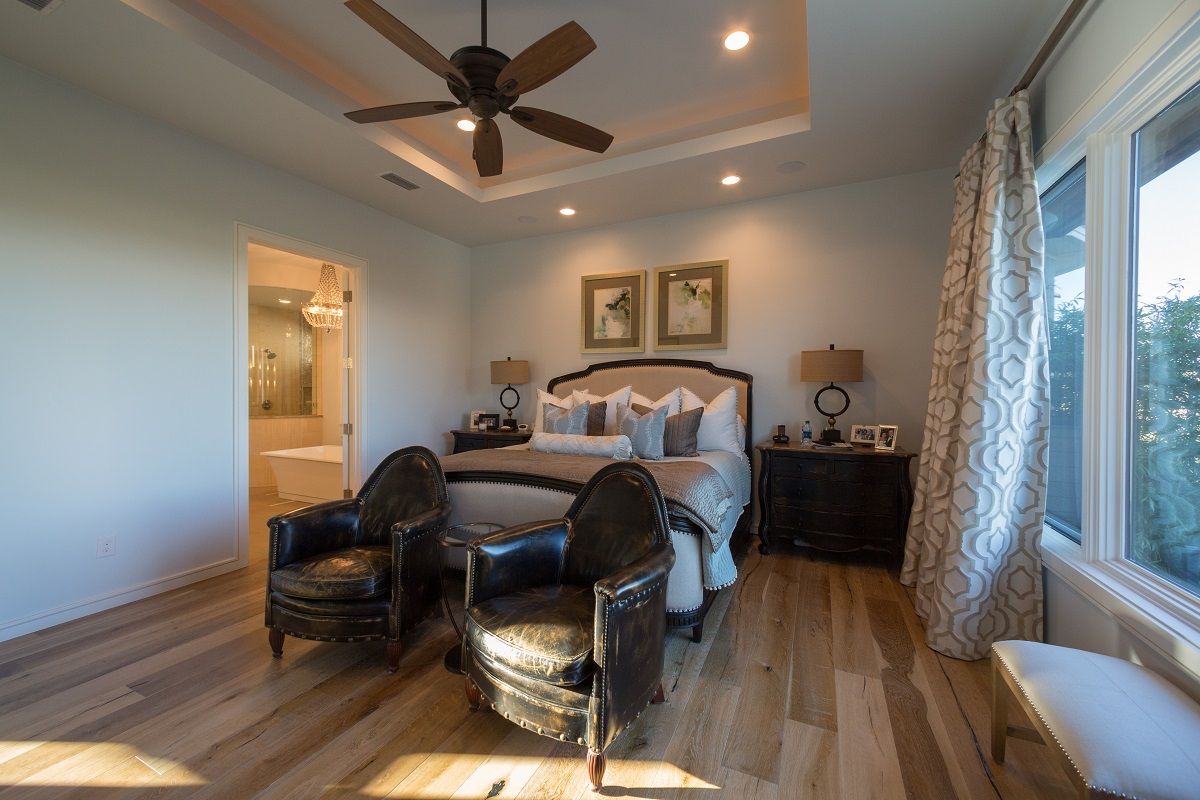 Bedroom with bed, chairs, and large window. Light wood floors, neutral walls, and decorative ceiling fan.