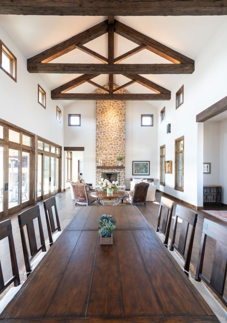 Long, rustic dining table in a room with high ceilings, wooden beams, and a stone fireplace.