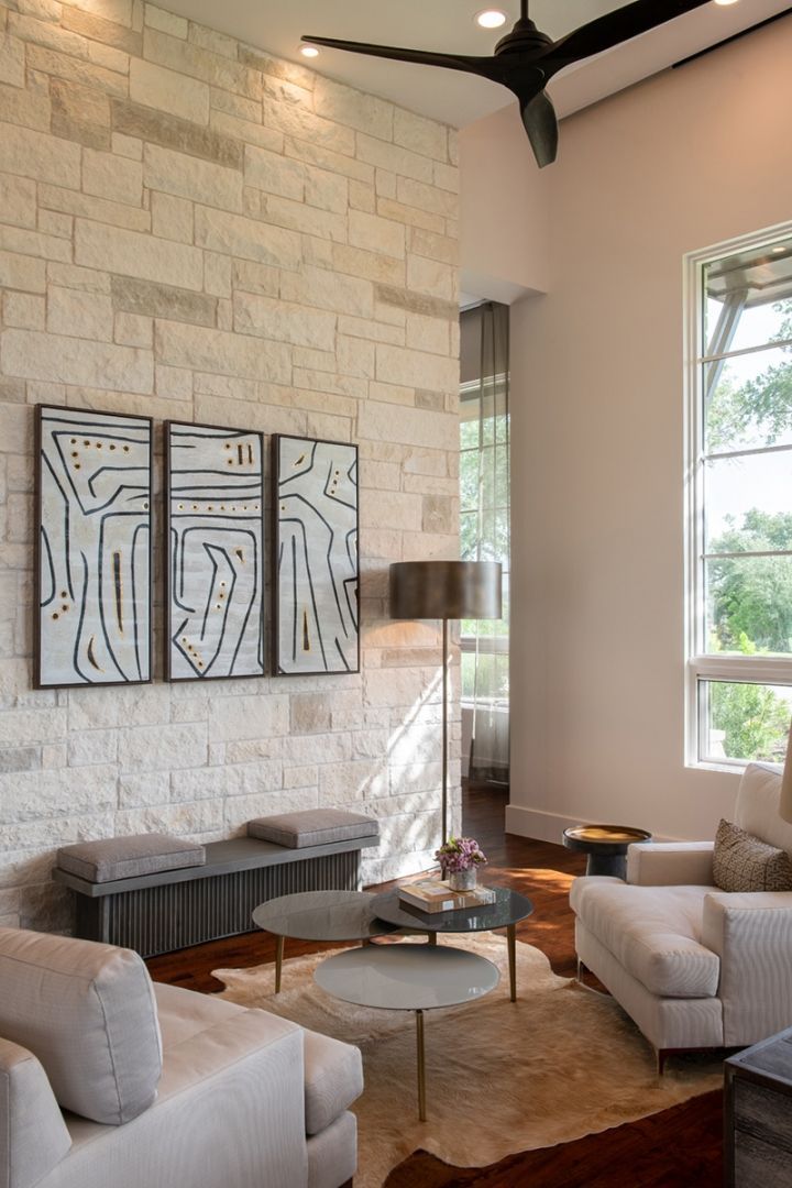 Living room with stone accent wall, abstract art, white sofas, and a view through a tall window.