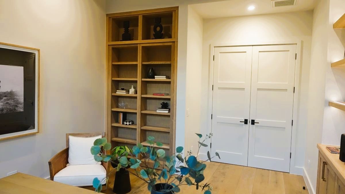 Office with built-in bookshelf, double doors, armchair, and desk; neutral tones with wooden and white accents.