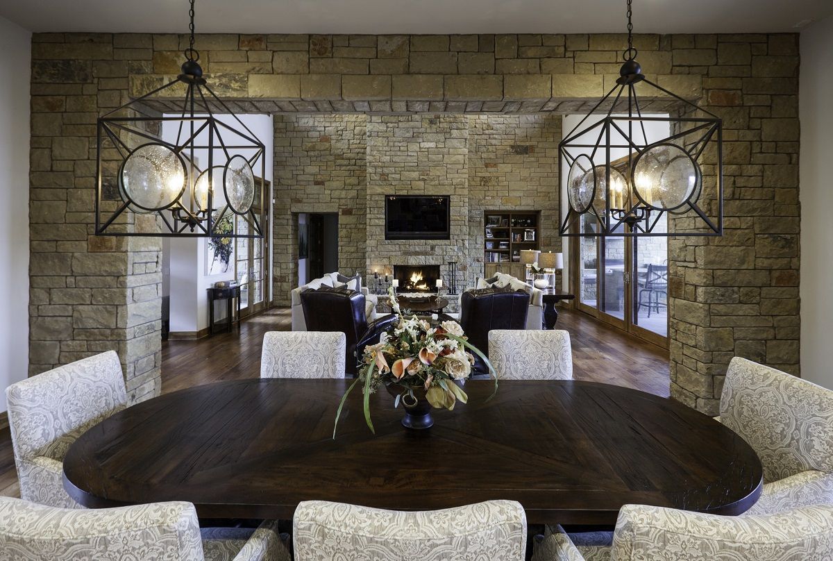 Formal dining room with oval table, stone walls, chandeliers, and view of living room.