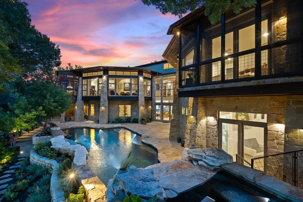 Luxury home with pool at sunset, stone exterior, large windows, trees, and a vibrant sky.