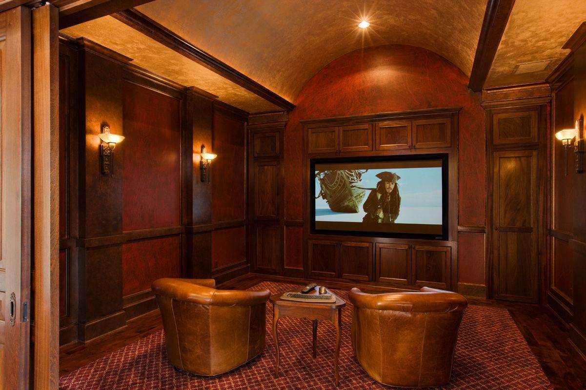 Cozy home theater with dark wood paneling, red walls, and leather chairs facing a large screen.