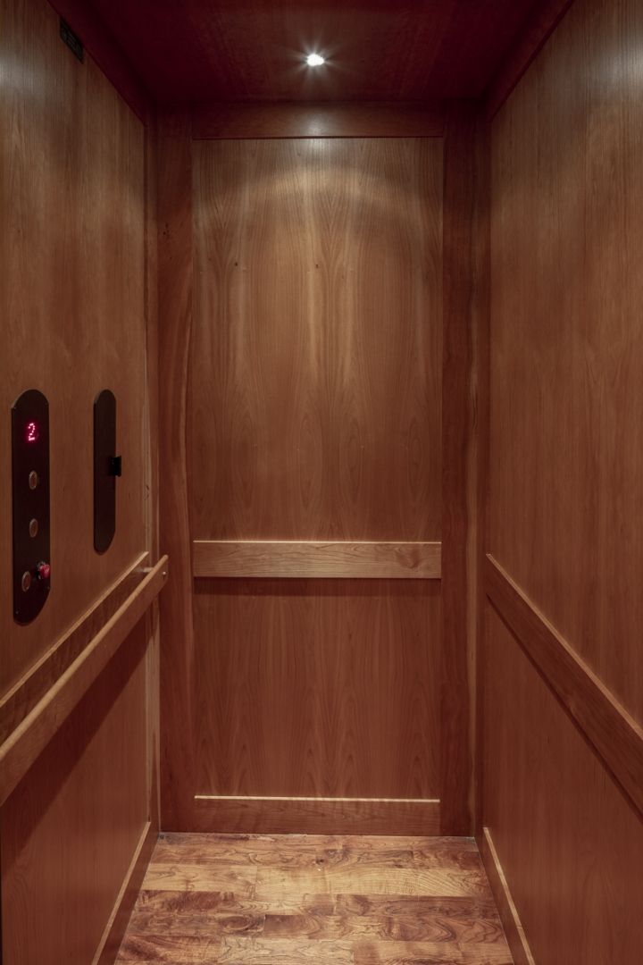 Wooden elevator interior, featuring door, control panel, and handrails. Warm tones of wood and a single ceiling light.