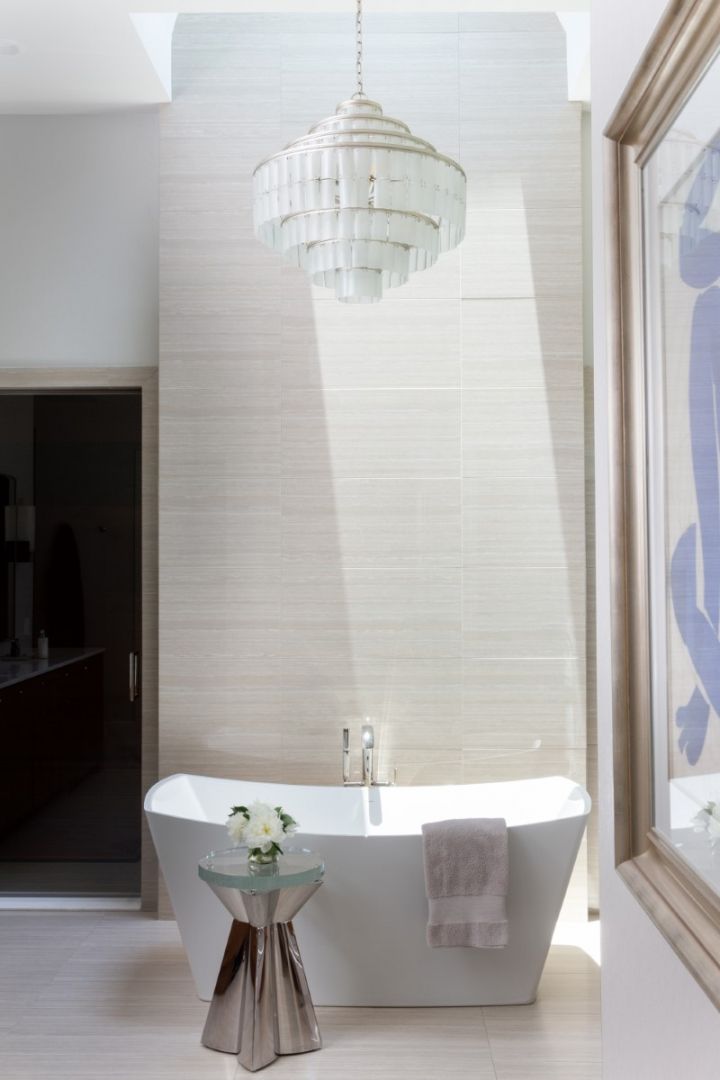 Bathroom with white tub, chandelier, silver side table, and light-colored wall and floor.