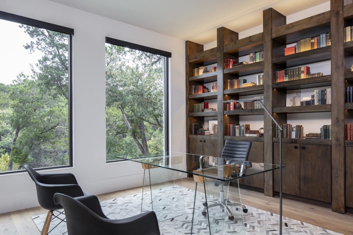 Modern home office with built-in bookshelves, glass desk, and black chairs, overlooking greenery.