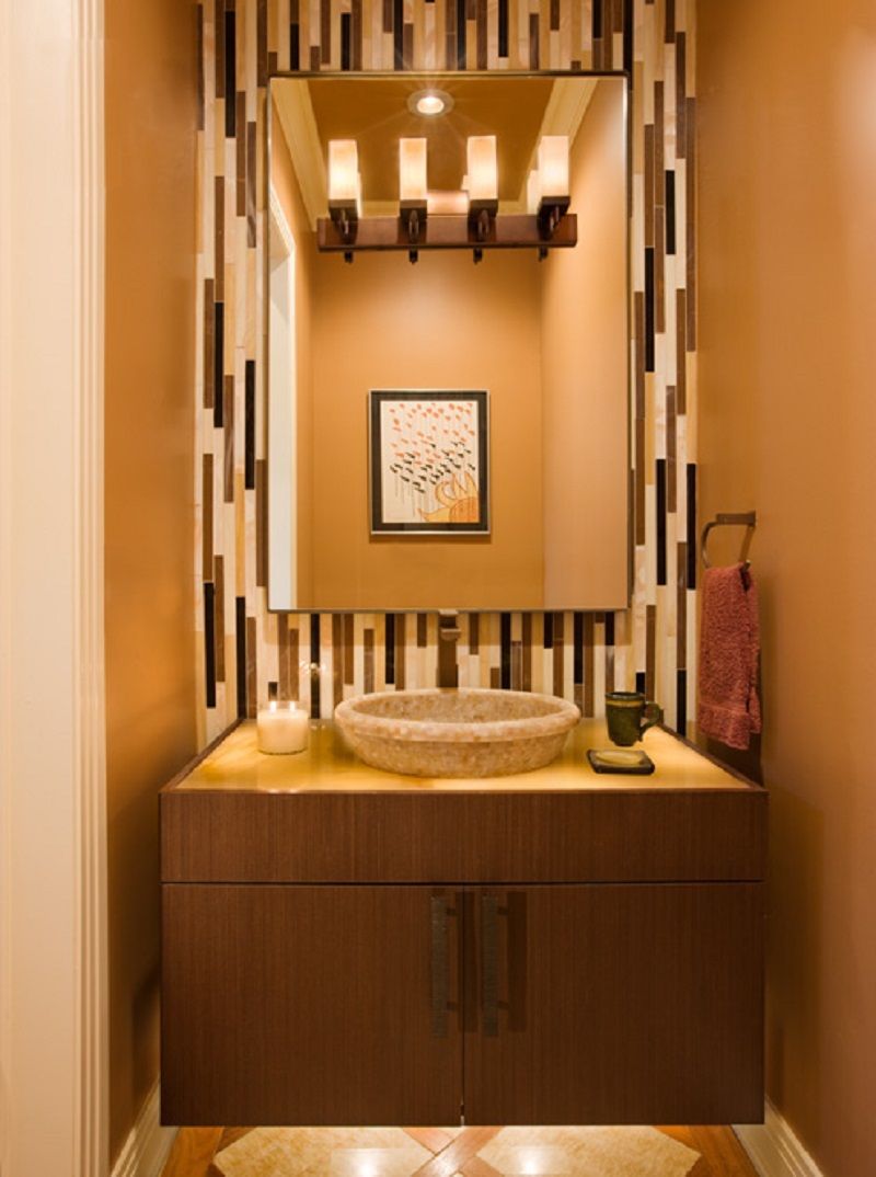 Small bathroom with brown cabinets, stone sink, mosaic accent wall, large mirror, and warm lighting.