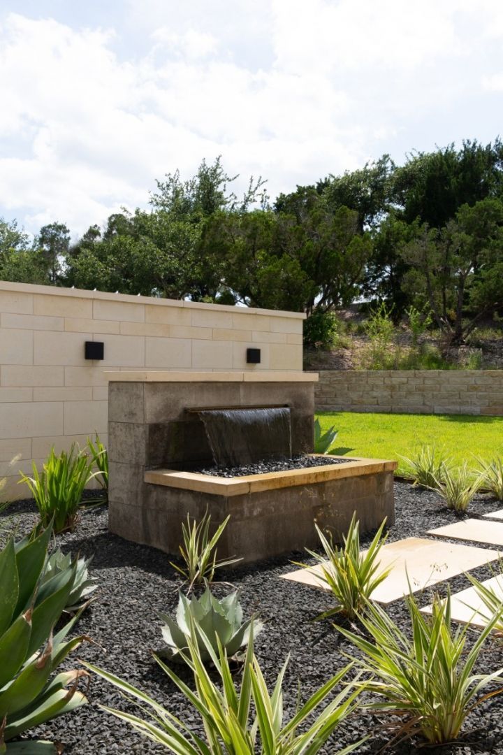 Fountain in a landscaped yard with black gravel and green plants; beige wall and trees in background.