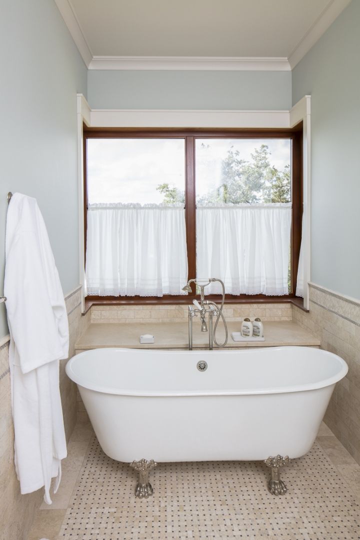 Clawfoot bathtub in a light blue bathroom, near a window with white curtains and a white robe.