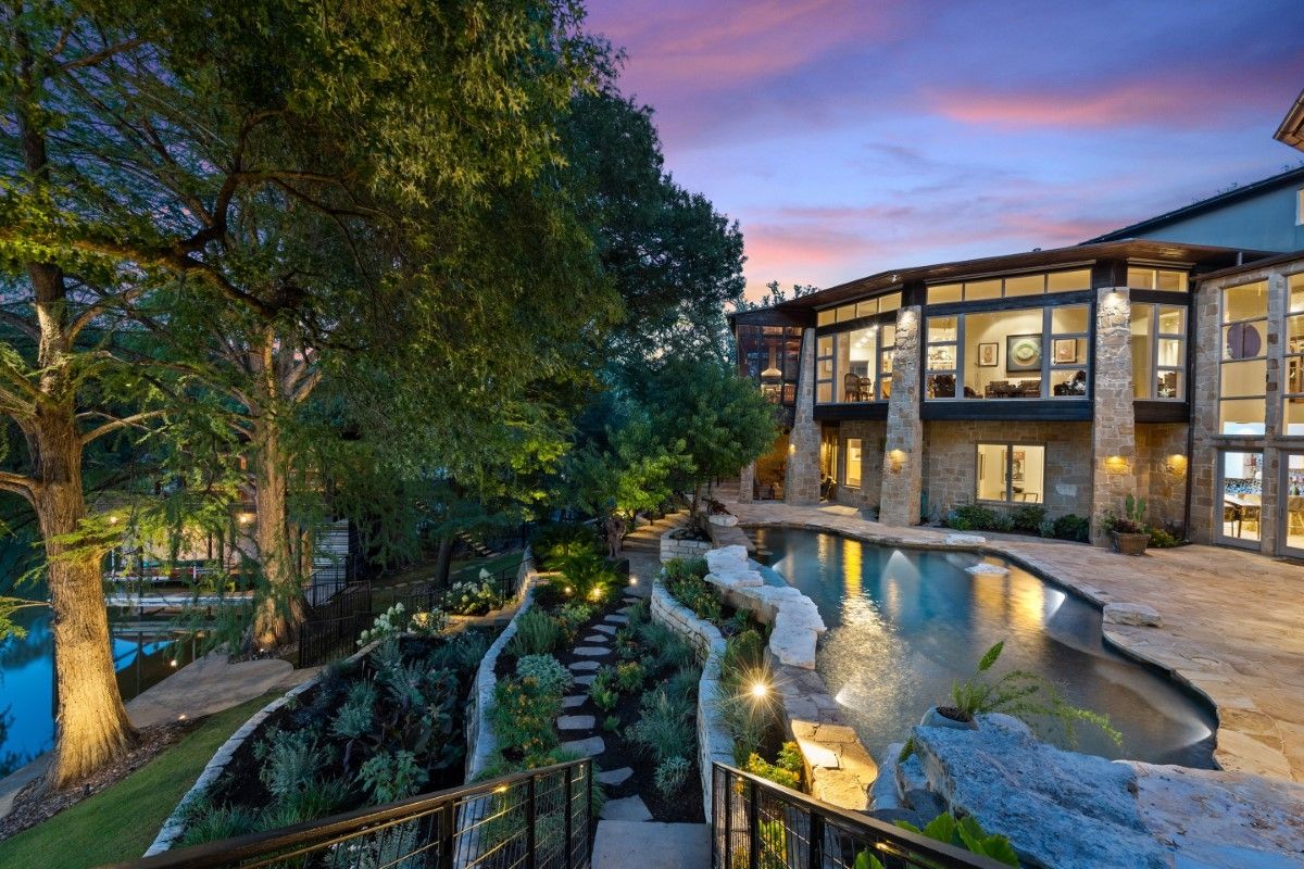 Luxury home with pool at dusk; landscaped grounds, stone steps, lake view, and vibrant sky.