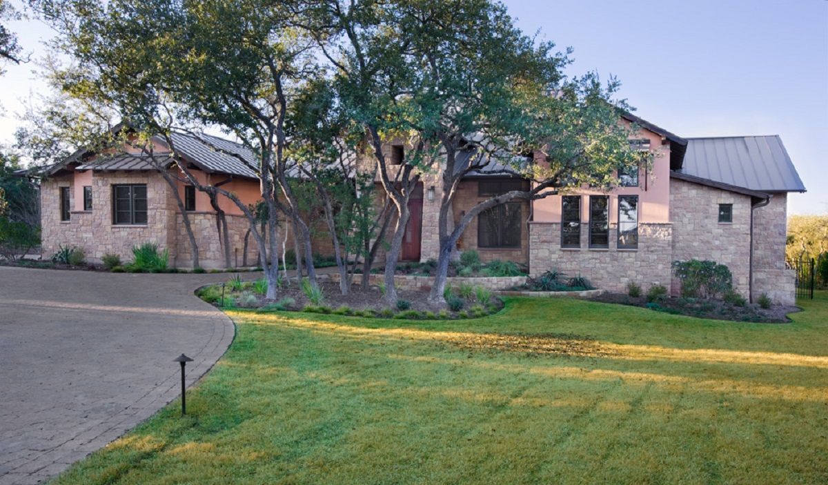Stone and stucco house with metal roof, set in a green yard with trees and driveway.