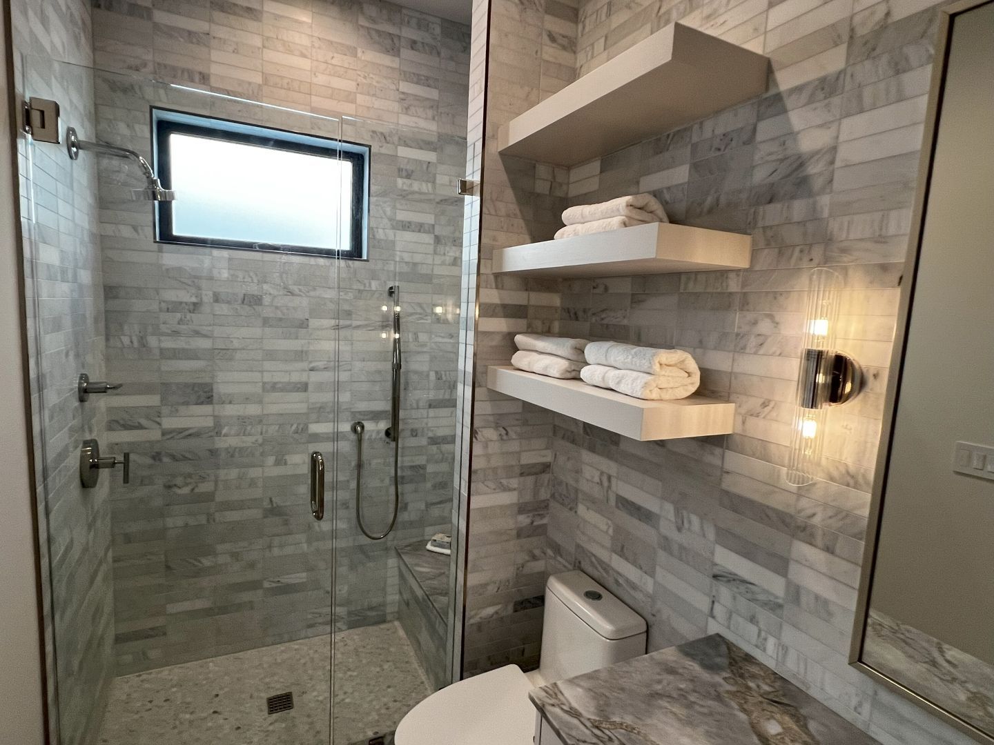 Small modern bathroom with a glass shower, white toilet, and marble-like tiled walls. Two shelves hold white towels.