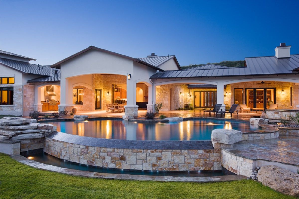 Luxury home at dusk with pool. Arched colonnades, stone facade, pool reflecting house lights.