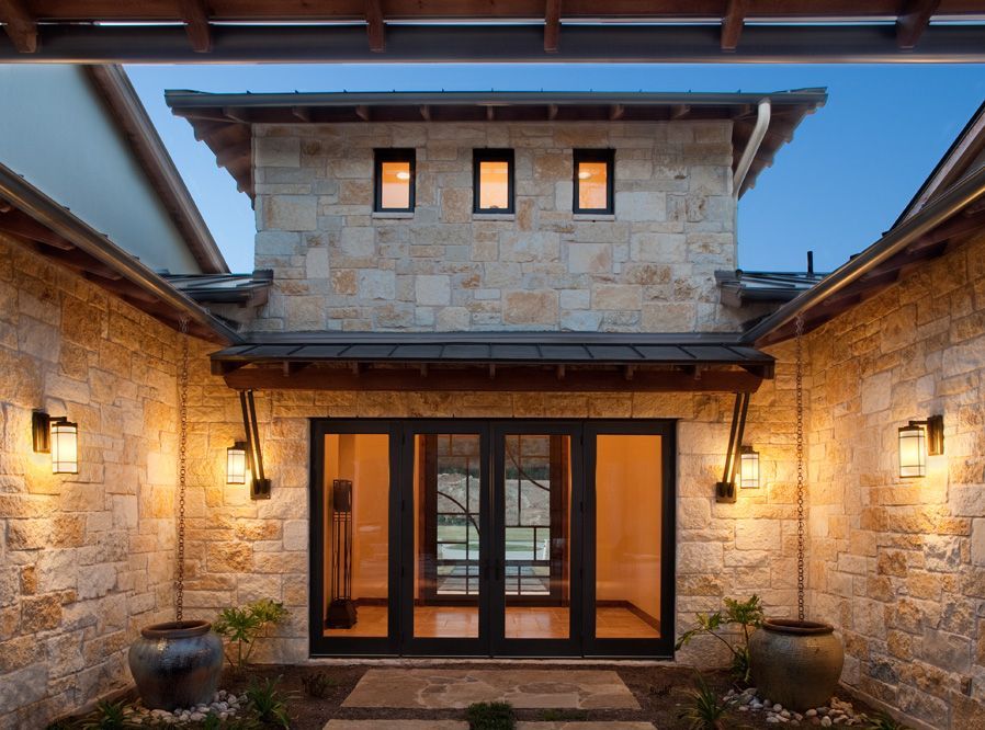 Stone house entrance with glass doors, sconce lights, and a small upper level with windows.