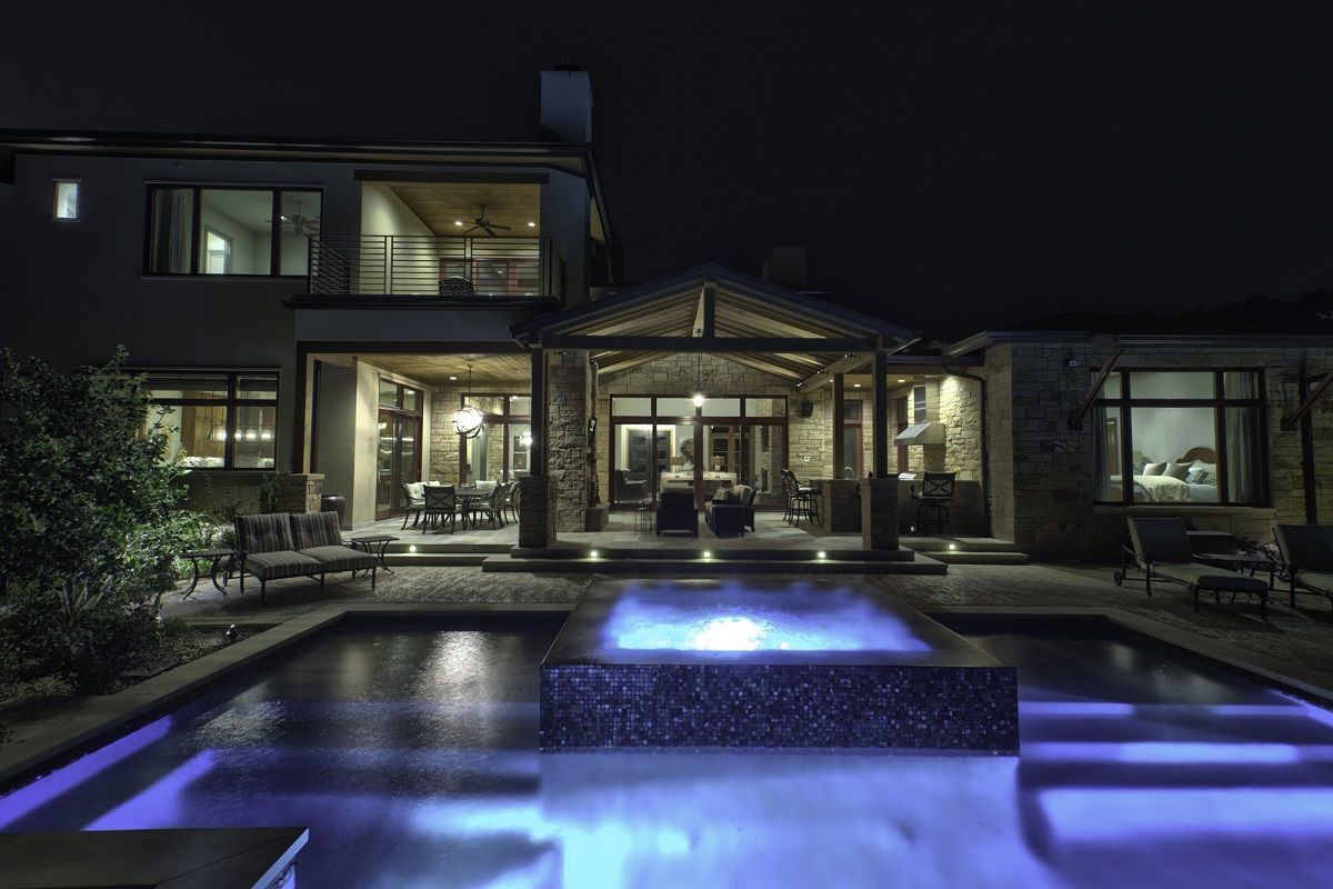 Nighttime view of a luxurious backyard with a pool. House lit up, with outdoor seating. Pool has blue lighting.