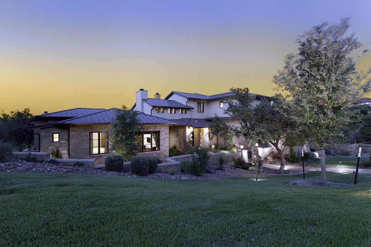 Stone house at dusk, with lit windows and landscaping; green lawn in foreground, blue sky.