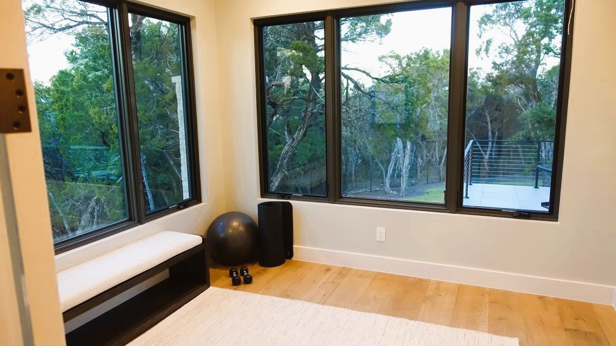 Home gym with large windows overlooking trees; bench, weights, and exercise ball.