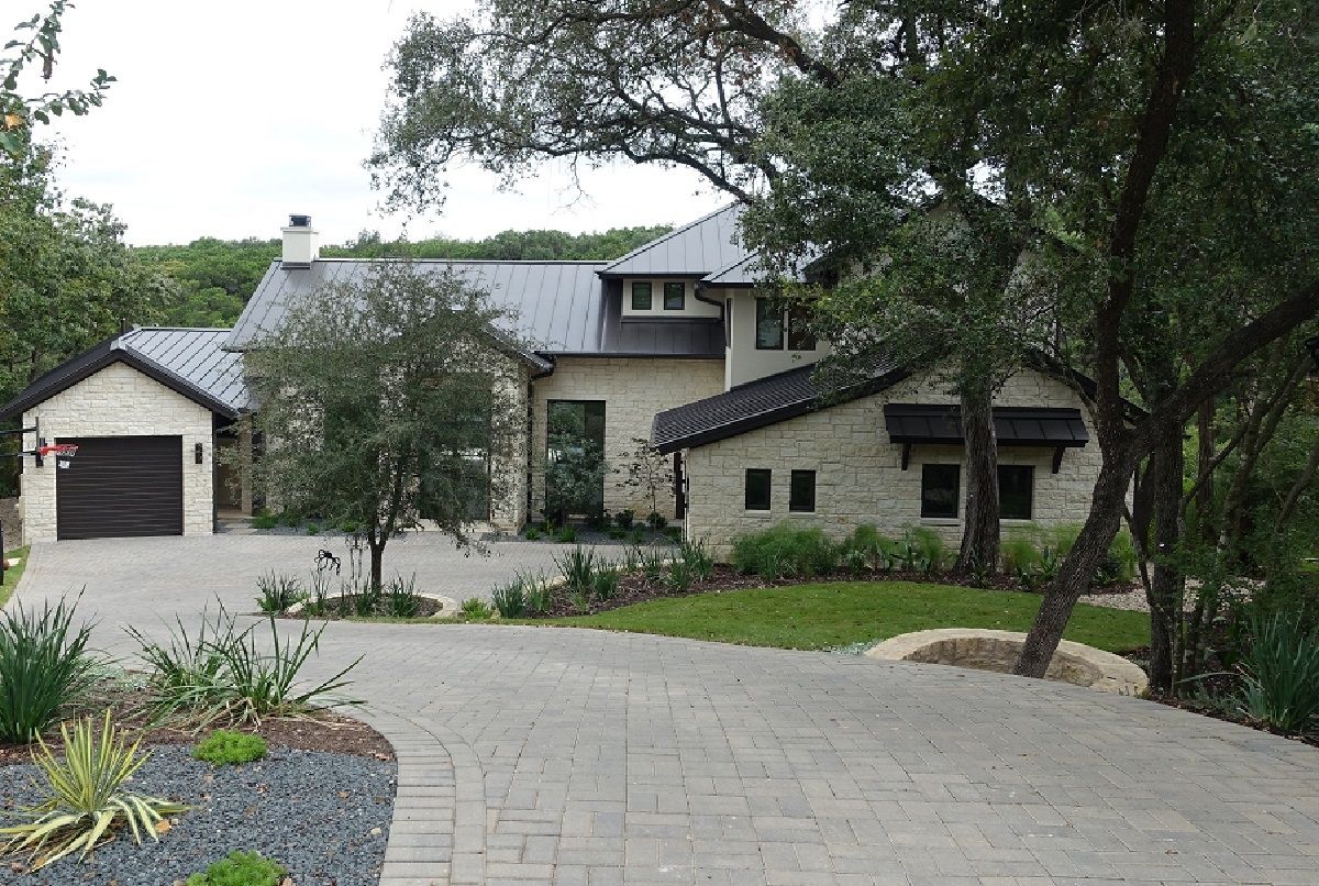 A modern, stone-clad house with a dark metal roof, driveway, and attached garage, nestled amongst trees.