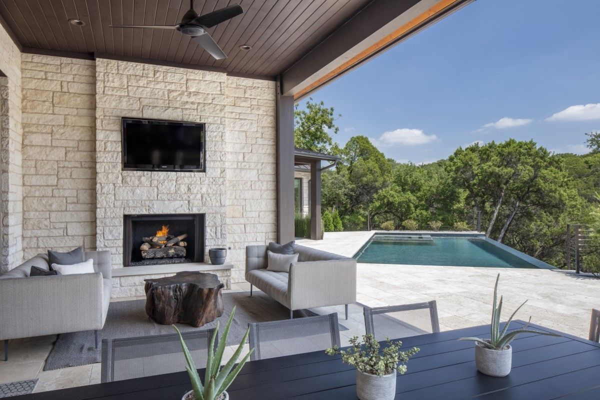Covered patio with fireplace, TV, and seating, overlooking a pool and trees.