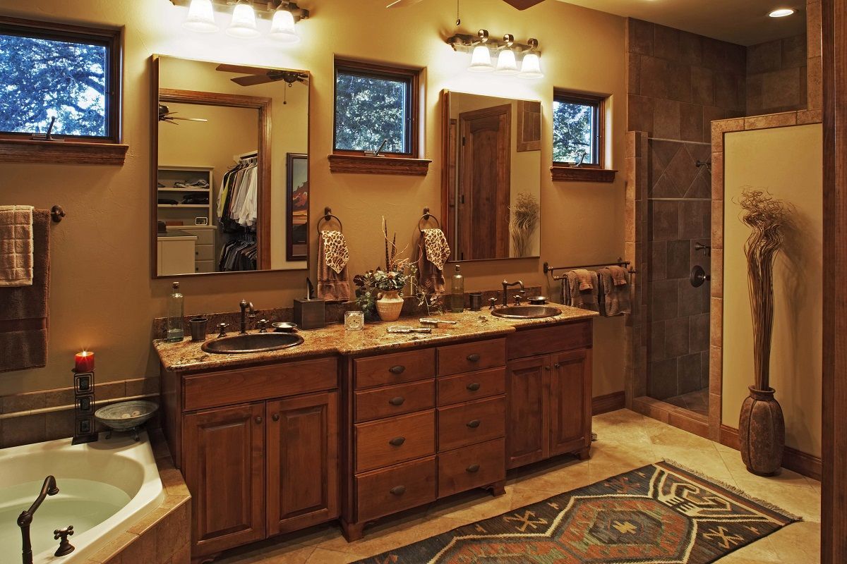 Bathroom with double vanity, mirrors, and a bathtub. Wooden cabinets, tan walls, and a patterned rug.