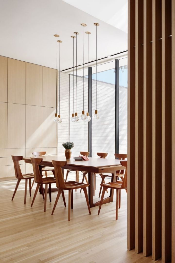 Dining room with wooden table, chairs, and hanging lights; light-colored walls and a slatted screen.
