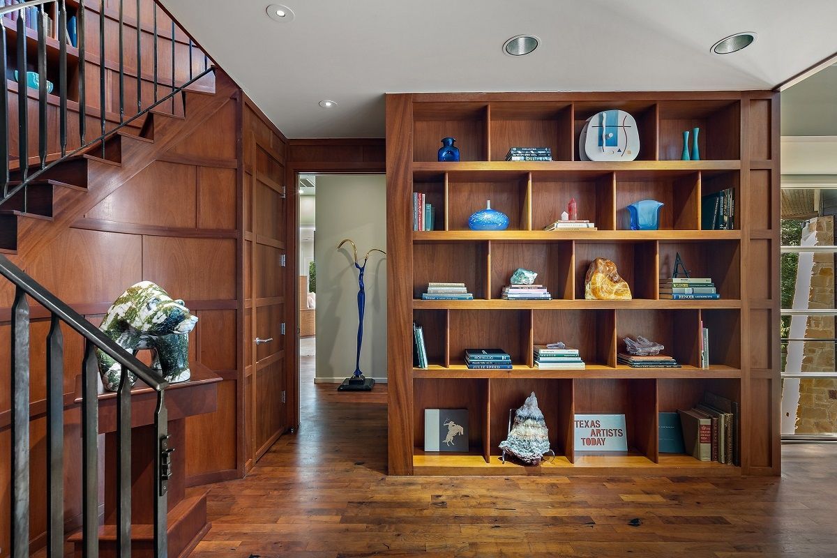 Wooden bookcase filled with objects in a hallway, beside a staircase with a metal railing.