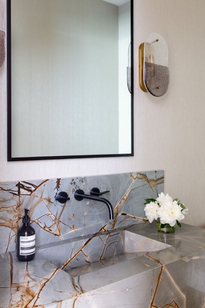 A modern bathroom with marble countertop and backsplash, black faucet, mirror, and a floral arrangement.