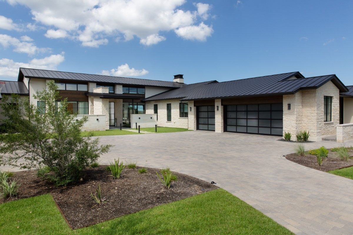 Modern two-story home with stone facade, black roof, glass garage doors, and concrete driveway under blue sky.