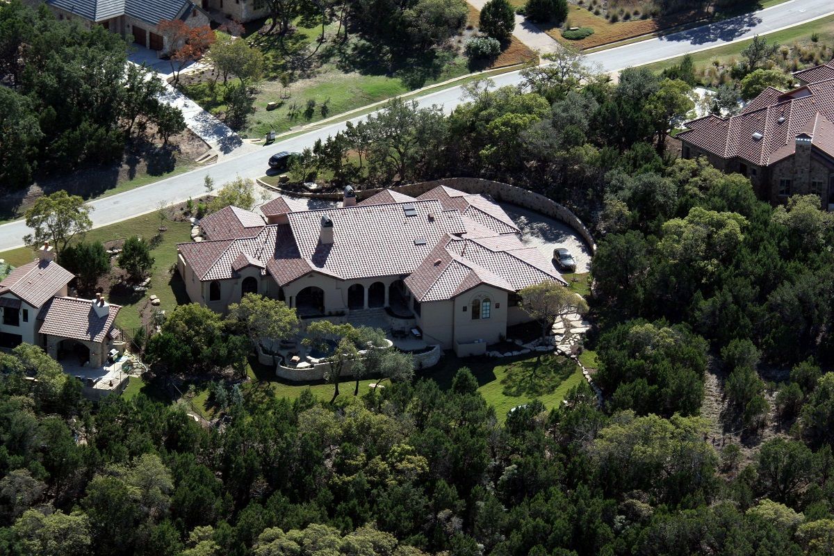 Aerial view of a large, tan-colored house with a curved roof and a circular driveway surrounded by trees.