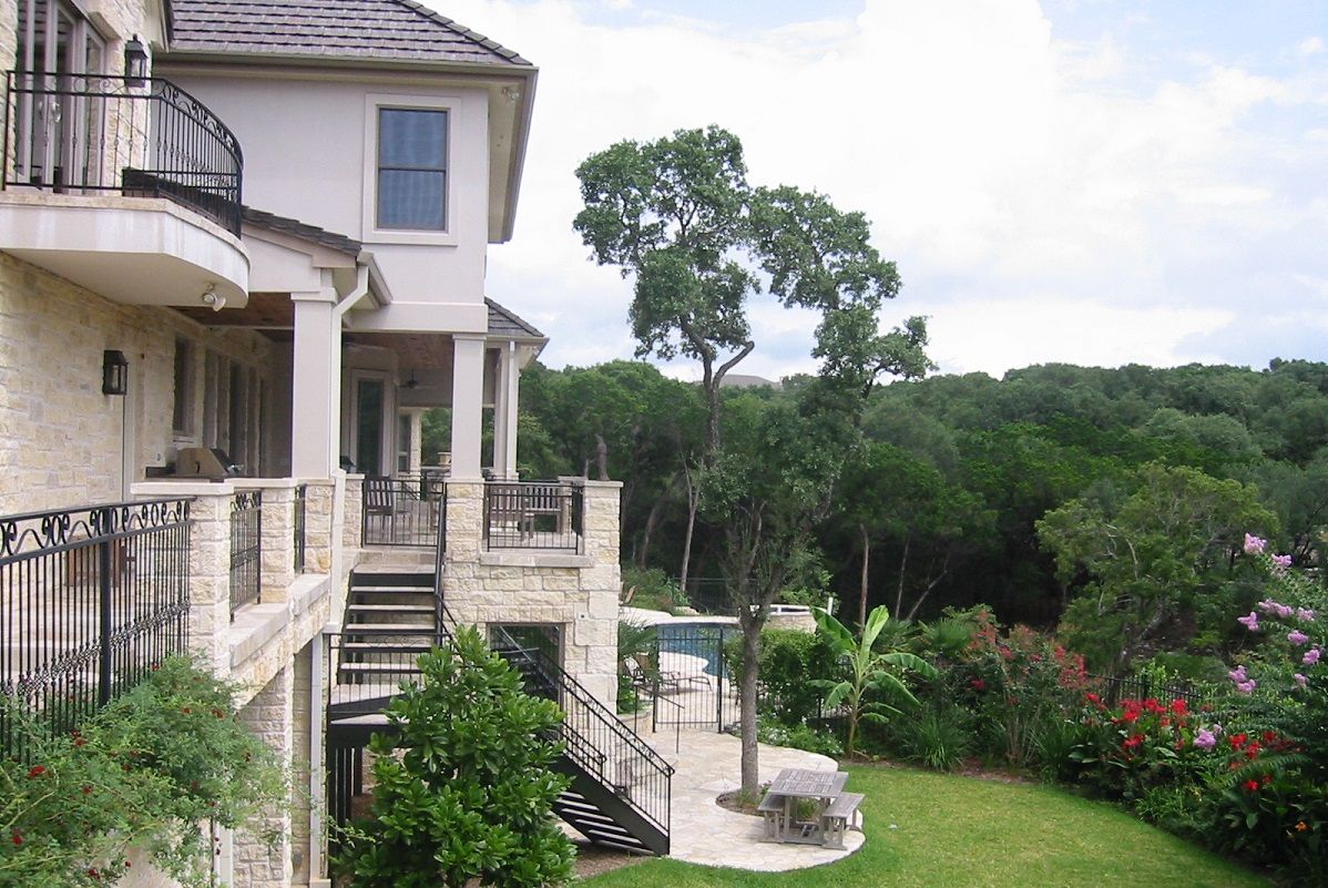 Two-story house with balcony overlooking a lush green yard, trees, and patio.