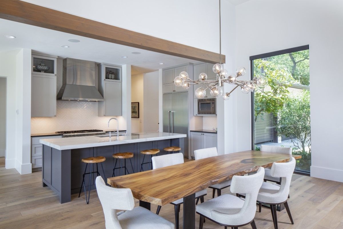 Modern kitchen and dining area with a dark gray island, wood dining table, and large window overlooking greenery.