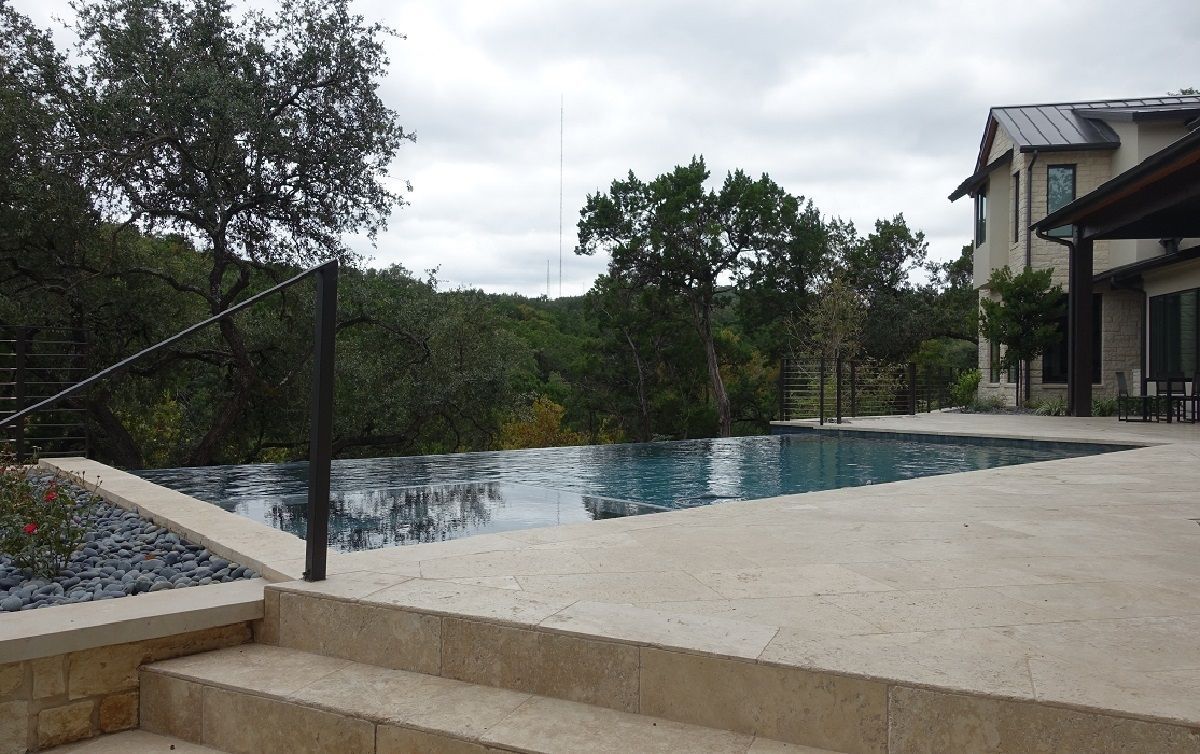 A pool with an infinity edge overlooks trees, next to a stone patio and house. Cloudy day.