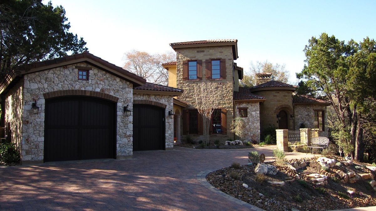 Stone-walled Mediterranean style home with a two-car garage, a tower, and brick driveway.
