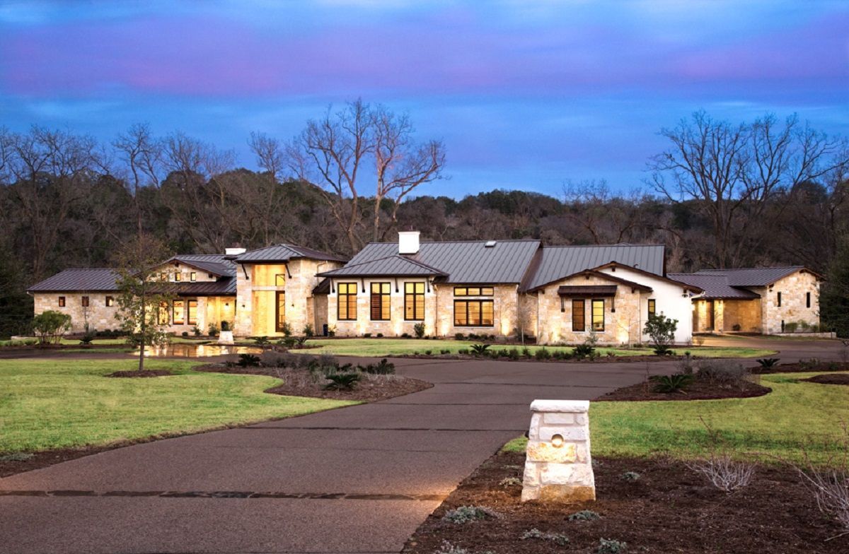 A modern, single-story home at dusk, with a long driveway, green lawn, and trees in the background.