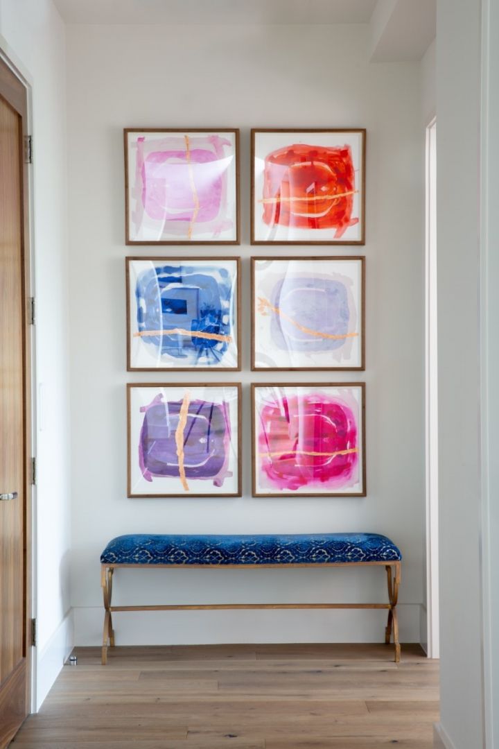 Six colorful abstract paintings above a blue bench in a hallway. Wooden door and white walls.