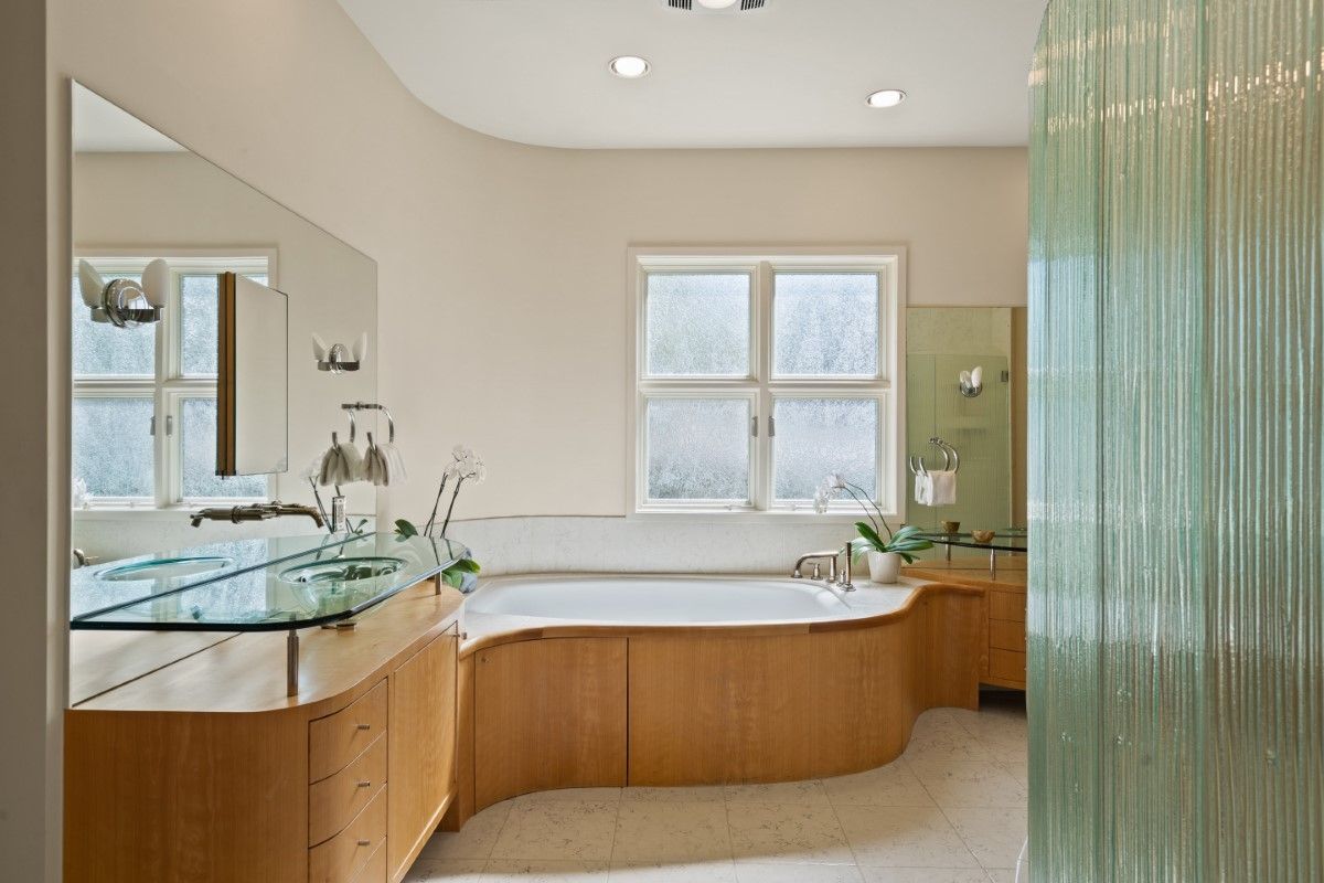 Modern bathroom with light wood cabinets, glass sink, and a curved bathtub under a window.