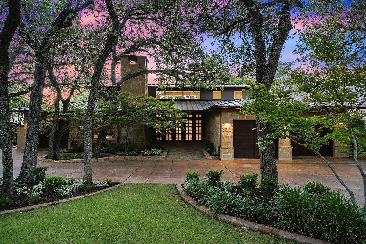 A modern home with stone facade and a lush green lawn, trees frame the entryway at sunset.