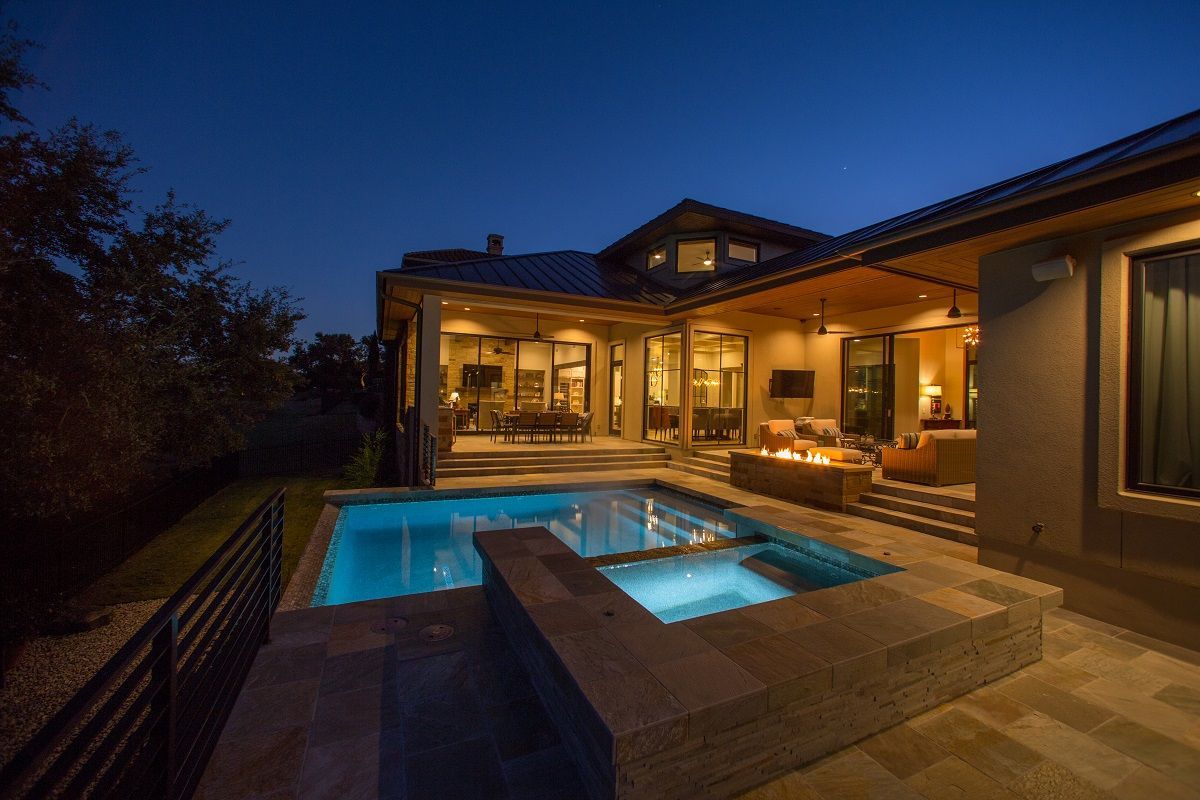 Night view of a luxury home with pool, spa, outdoor seating, and fire pit; blue water, warm lighting.