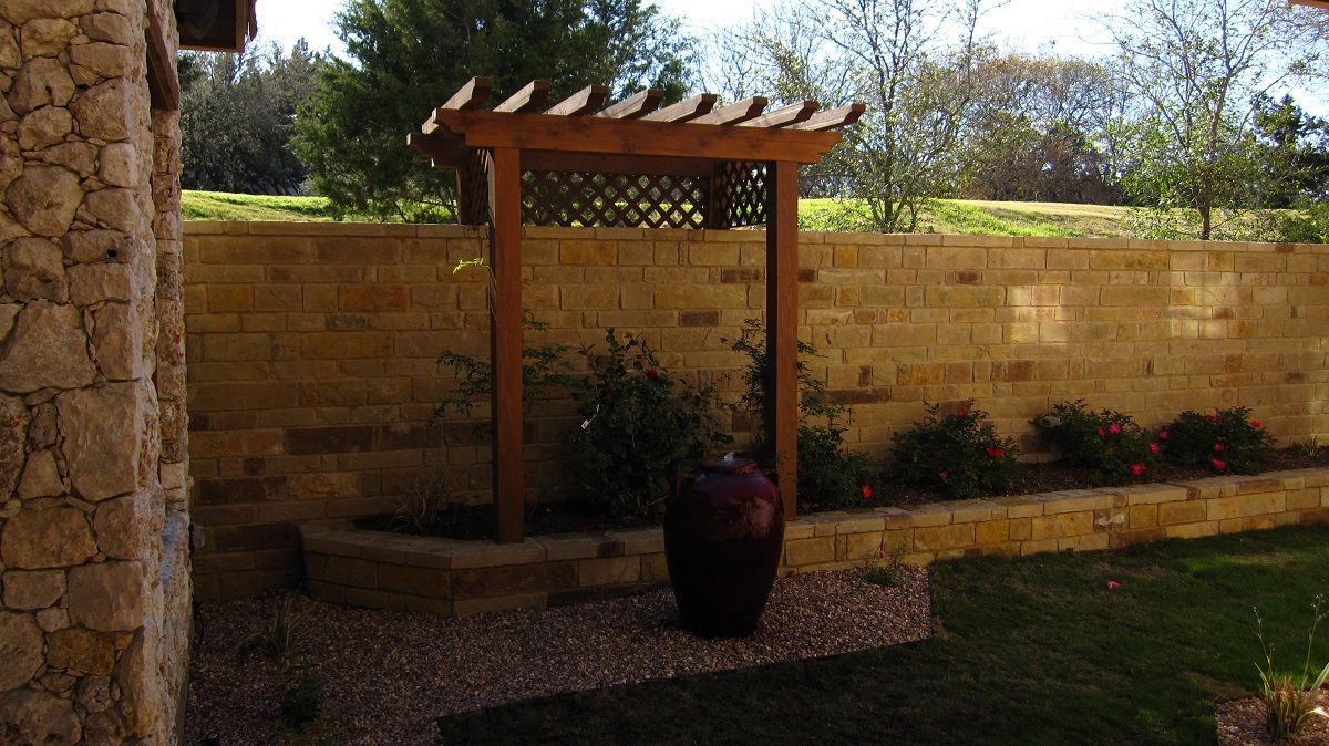 Wooden arbor in a garden with stone walls, a large pot, and green grass.