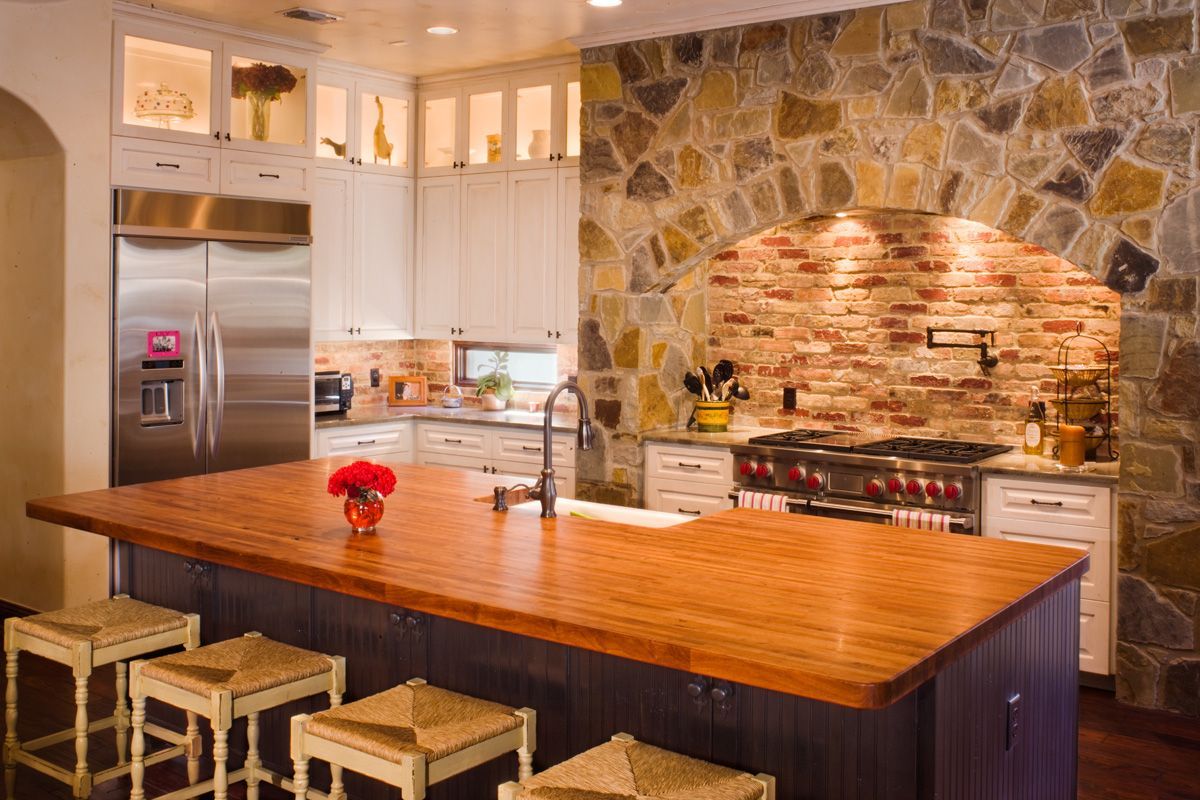 Kitchen with a large wooden island and a brick oven, light cabinetry and stone wall.
