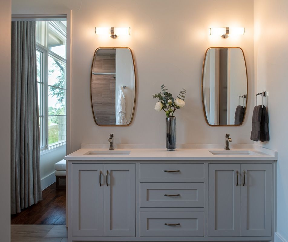 Gray bathroom vanity with two sinks, mirrors, and sconces, flowers, and towels.