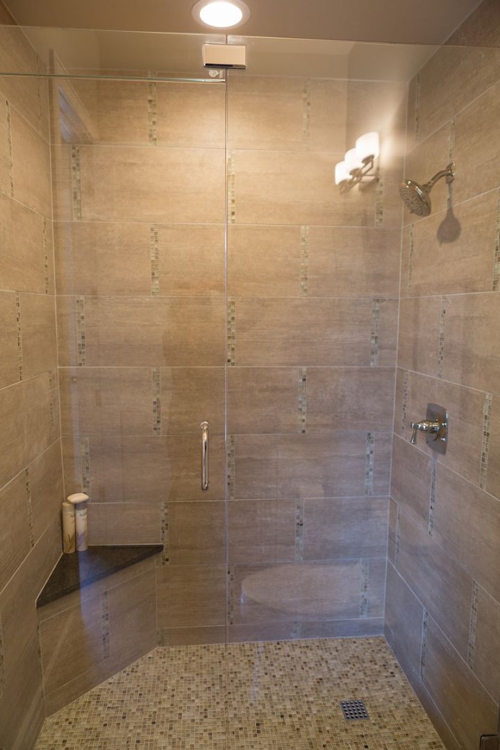 Shower with beige tile walls, pebble floor, glass door and corner shelf.