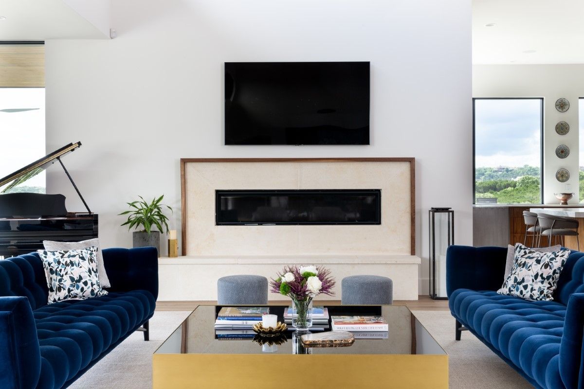 Living room with blue velvet sofas, a grand piano, fireplace, and large TV on a white wall.